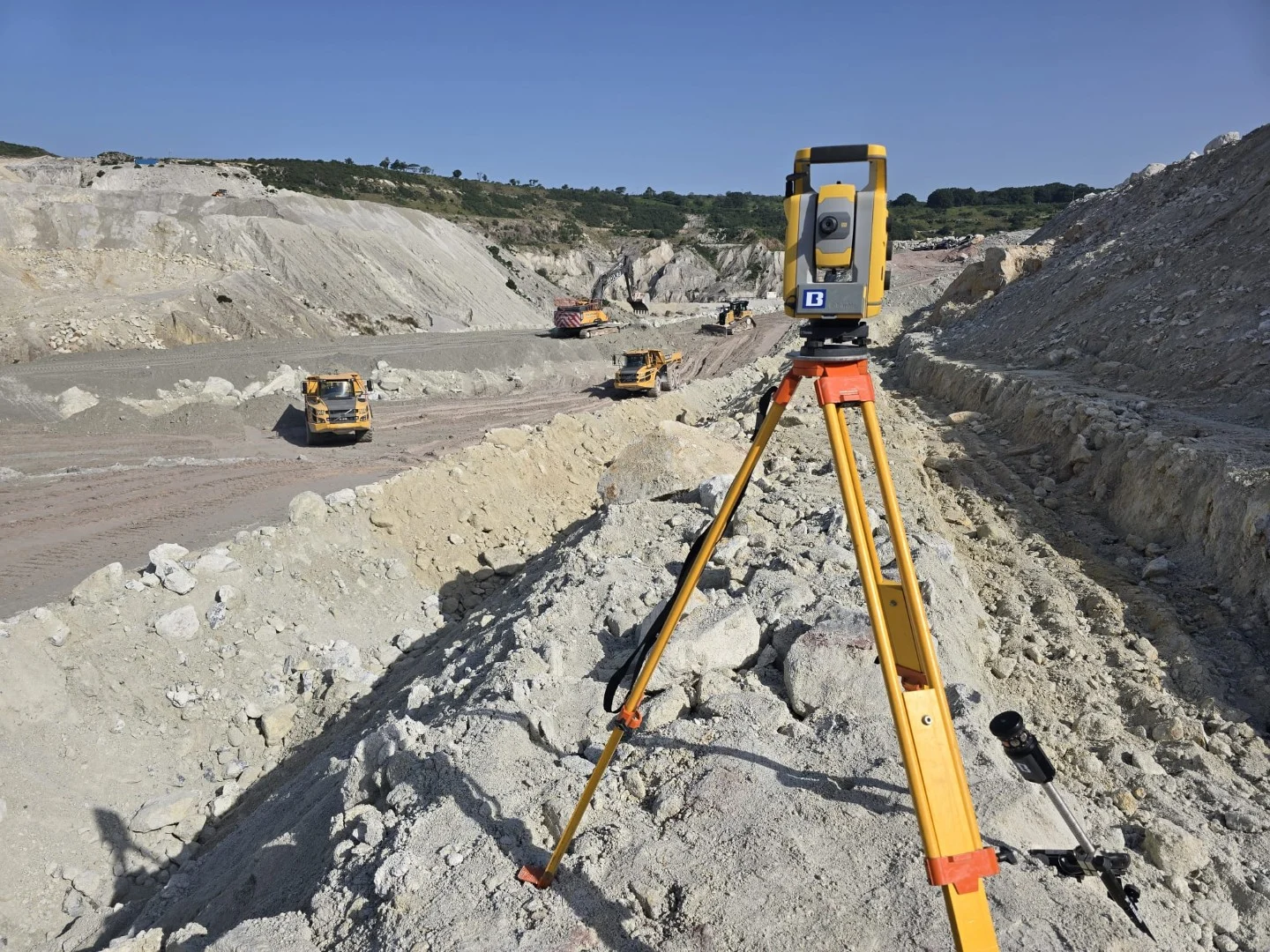 Total station on a tripod overlooking an excavated site with haul roads and construction vehicles