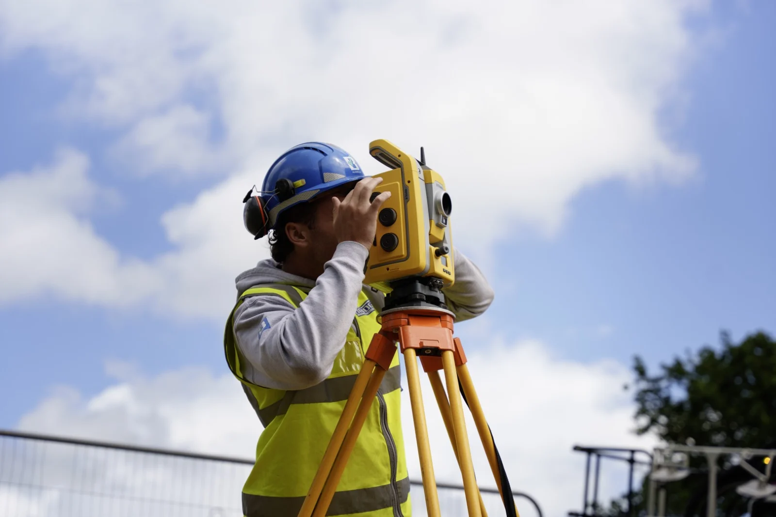 Surveyor in a blue hard hat and high-visibility vest using a total station on a tripod