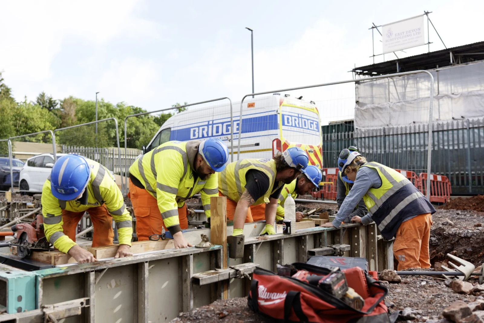 Bridge construction team in hard hats and high-visibility clothing working on formwork beside a company van