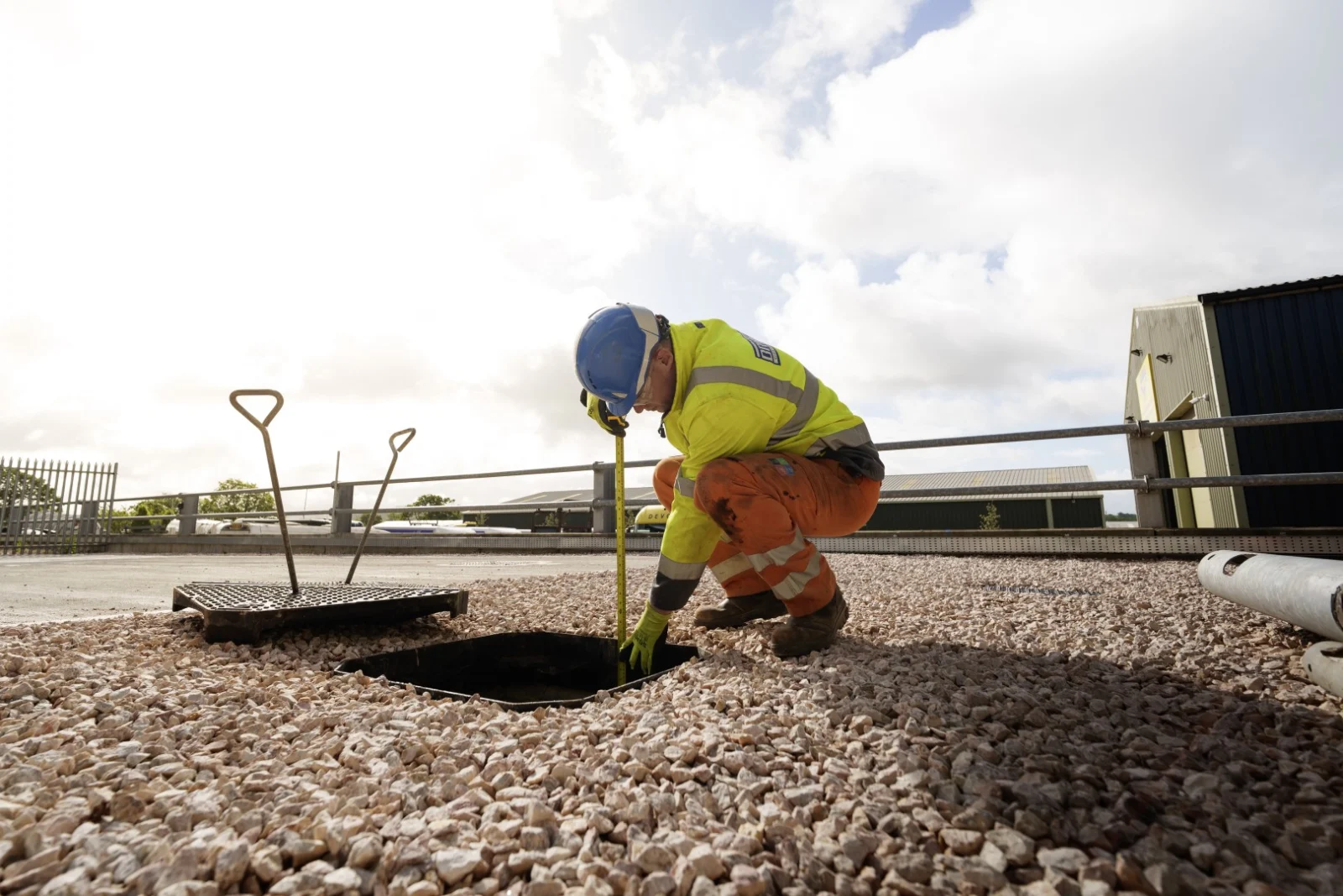 Worker in high-visibility clothing measuring inside an open access chamber in a gravel surface