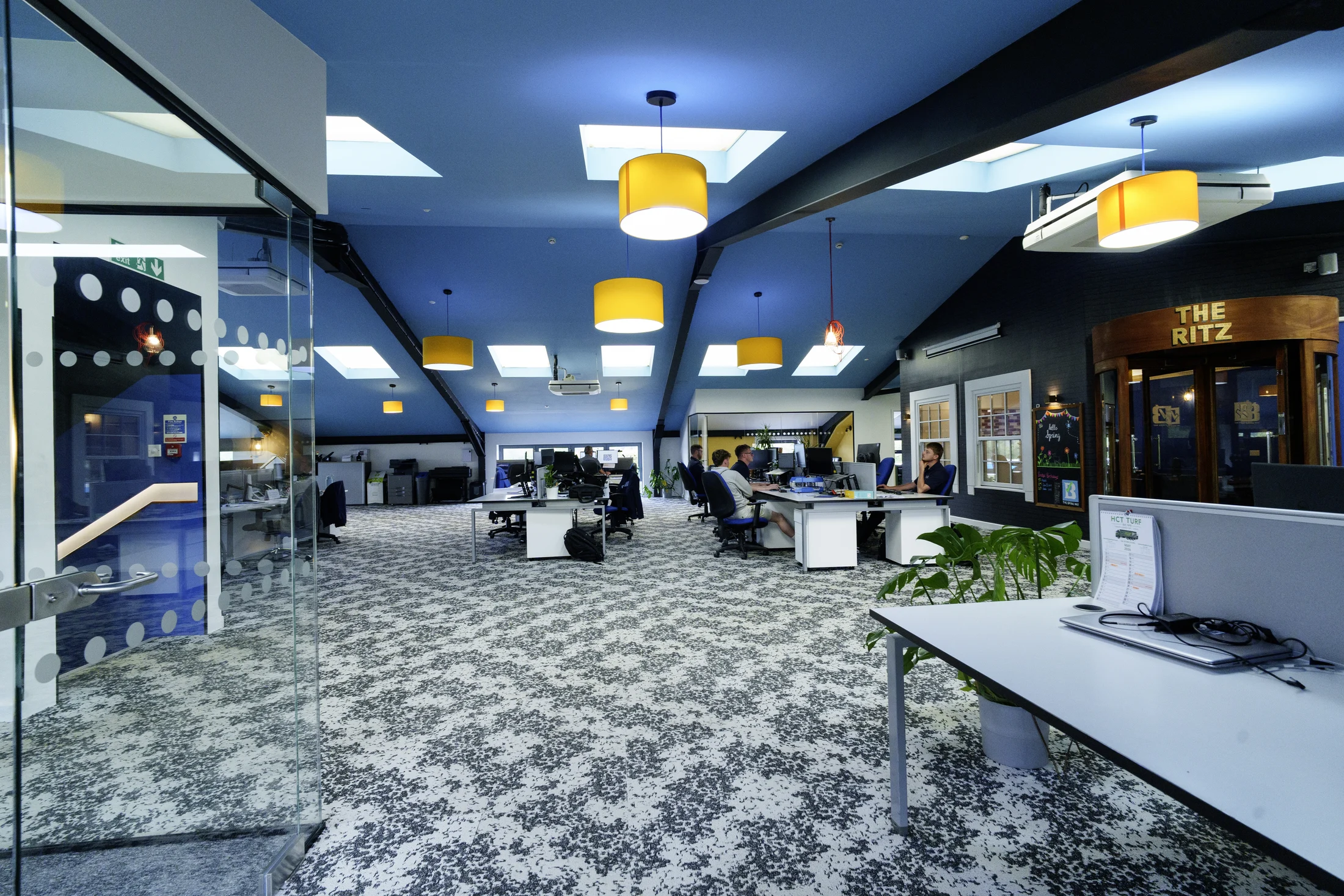 Open-plan Bridge office with colleagues at desks, glass meeting rooms, blue ceilings and yellow pendant lights
