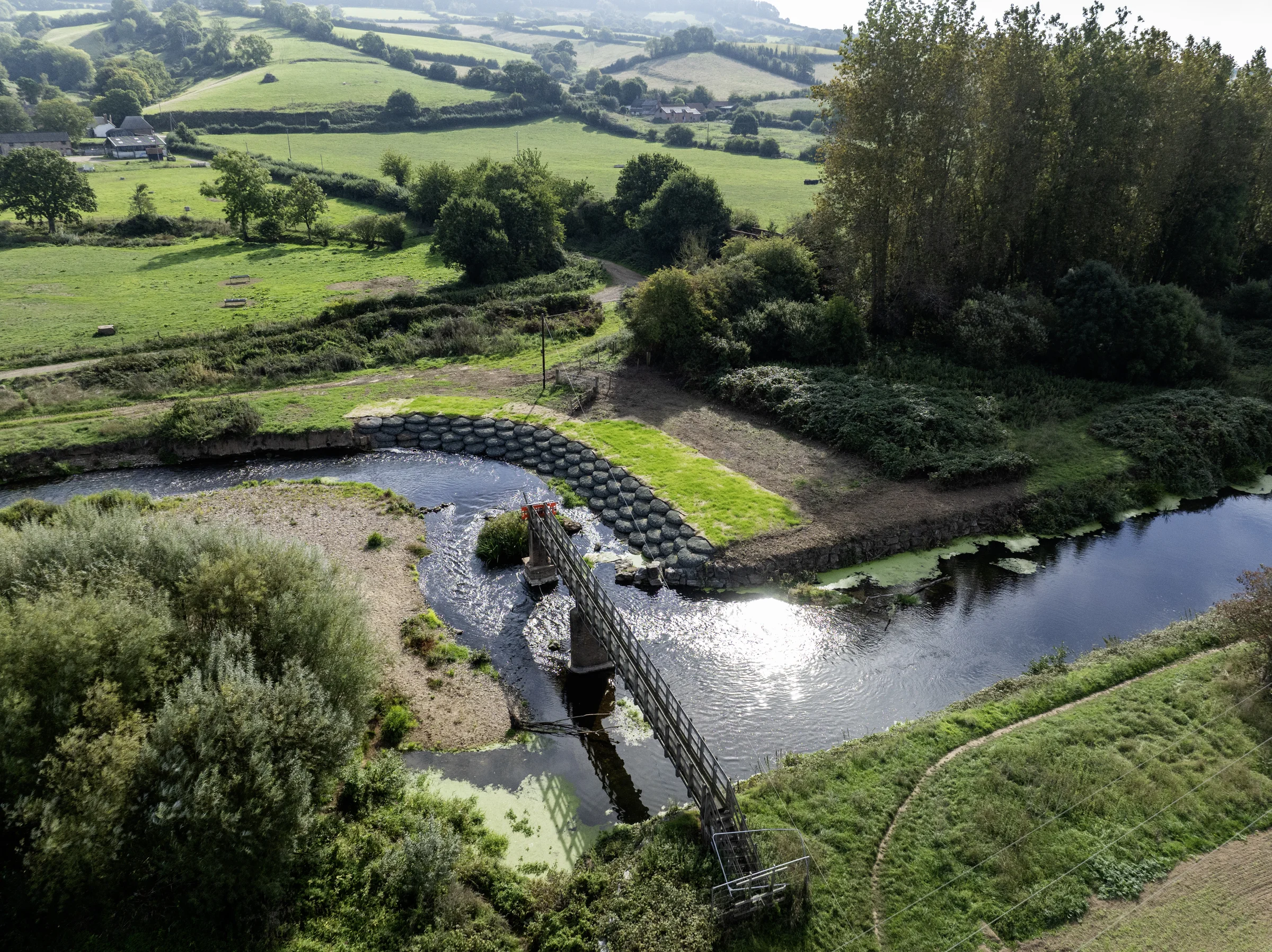 Aerial view of a river weir and footbridge surrounded by trees and green countryside