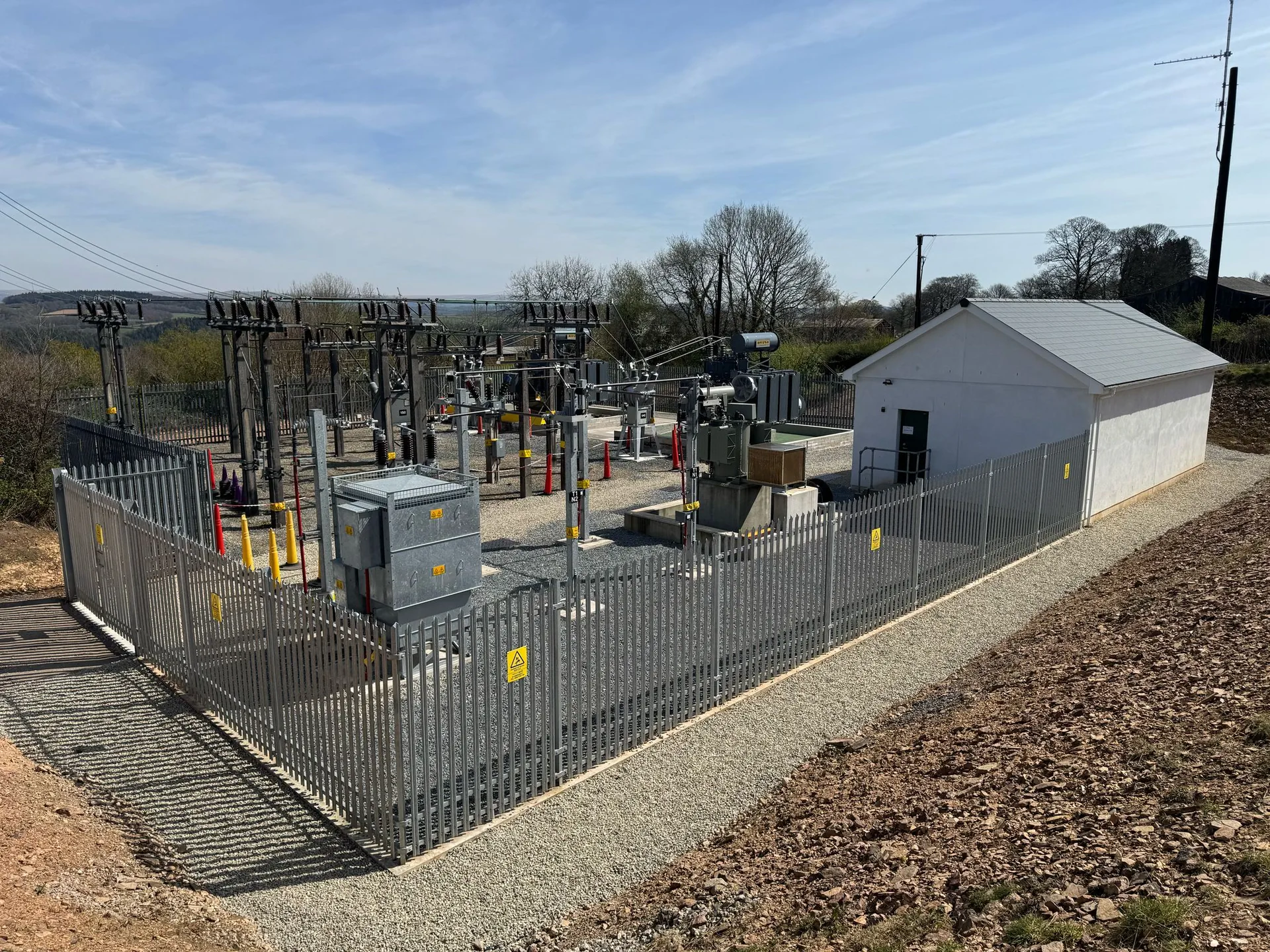 Electrical substation compound with switchgear behind steel palisade fencing, beside a single-storey control building