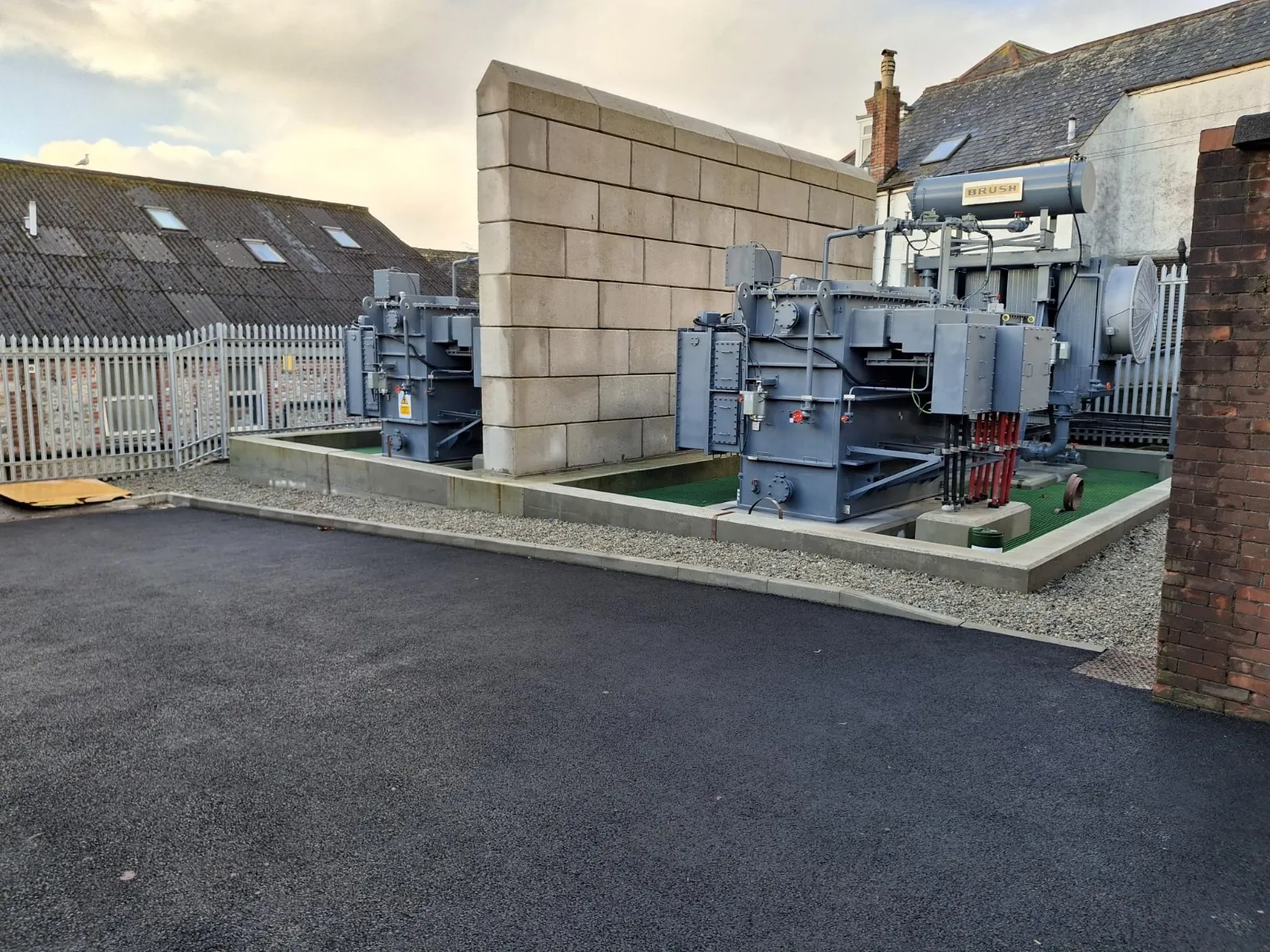 Substation transformers inside a walled enclosure next to a freshly surfaced tarmac yard