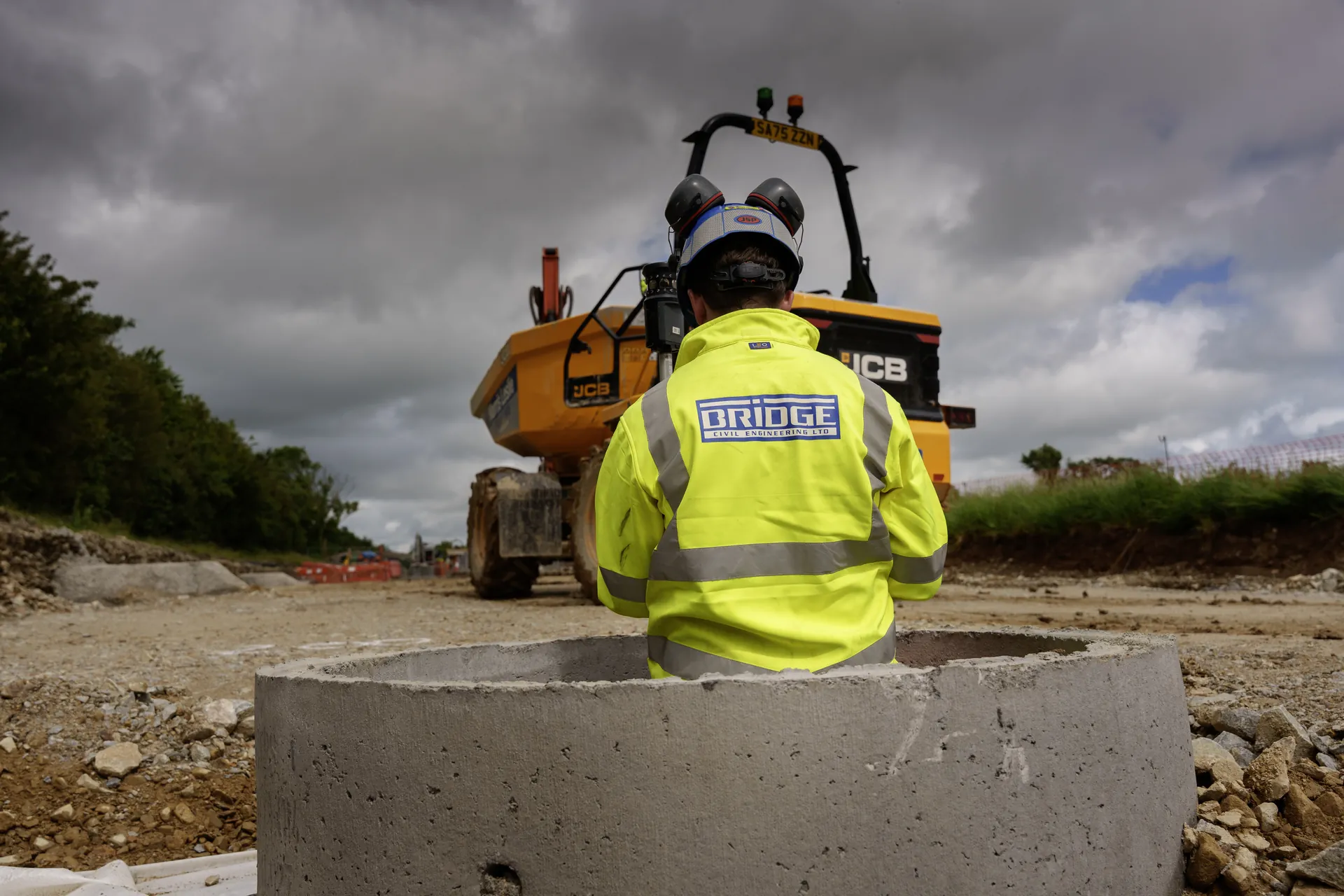 Operative in a Bridge hi-vis jacket and hard hat behind a precast concrete chamber ring, with a JCB dumper beyond