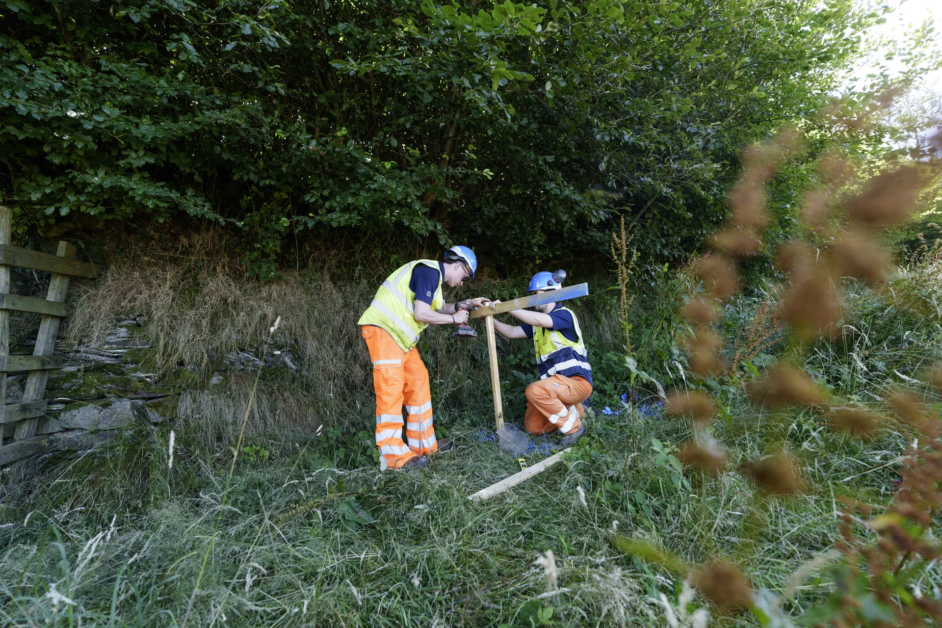 Two operatives in high-visibility clothing working on a timber setting-out frame beside a hedgerow
