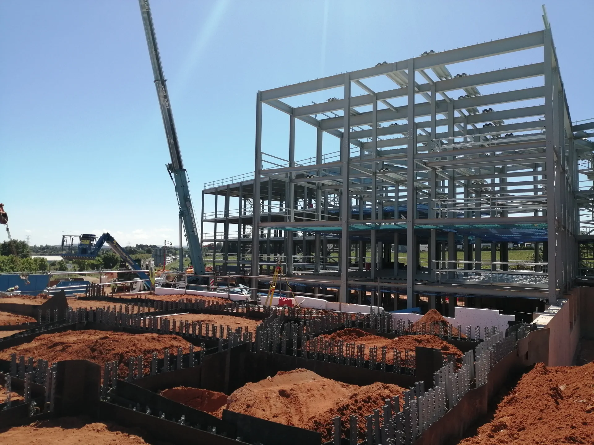 Multi-storey steel frame under construction, with a crane and foundation works with projecting reinforcement in the foreground