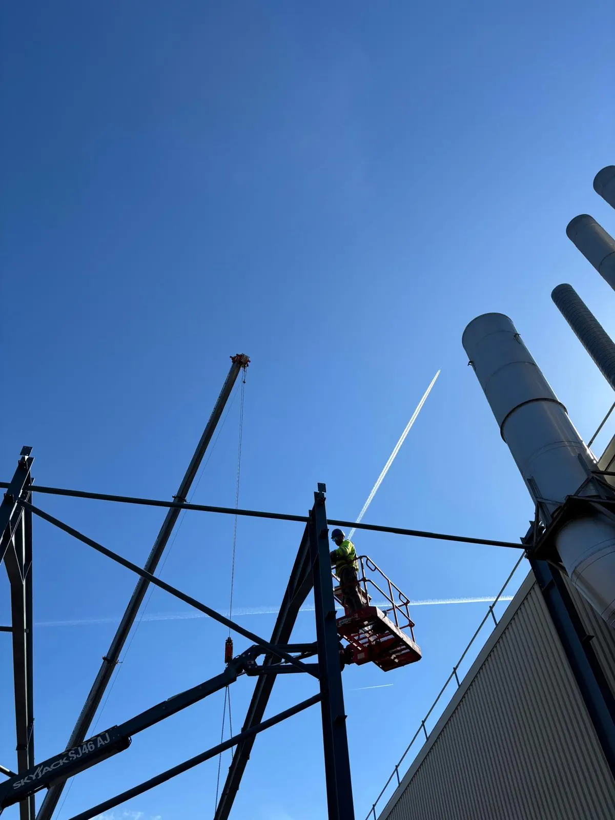 Operative in a raised work platform beside a steel frame and tall industrial stacks against a blue sky