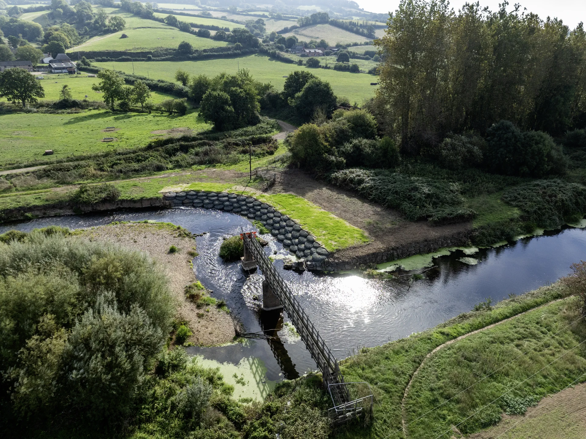 Aerial view of a river with an angled in-river structure, rock-lined banks and surrounding fields and trees