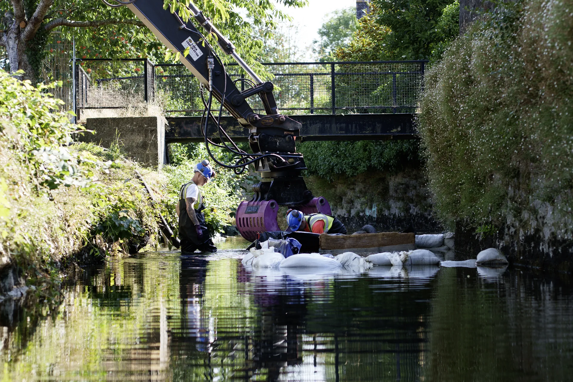 Operatives working in a river beside an excavator arm and a line of sandbags, with a footbridge behind