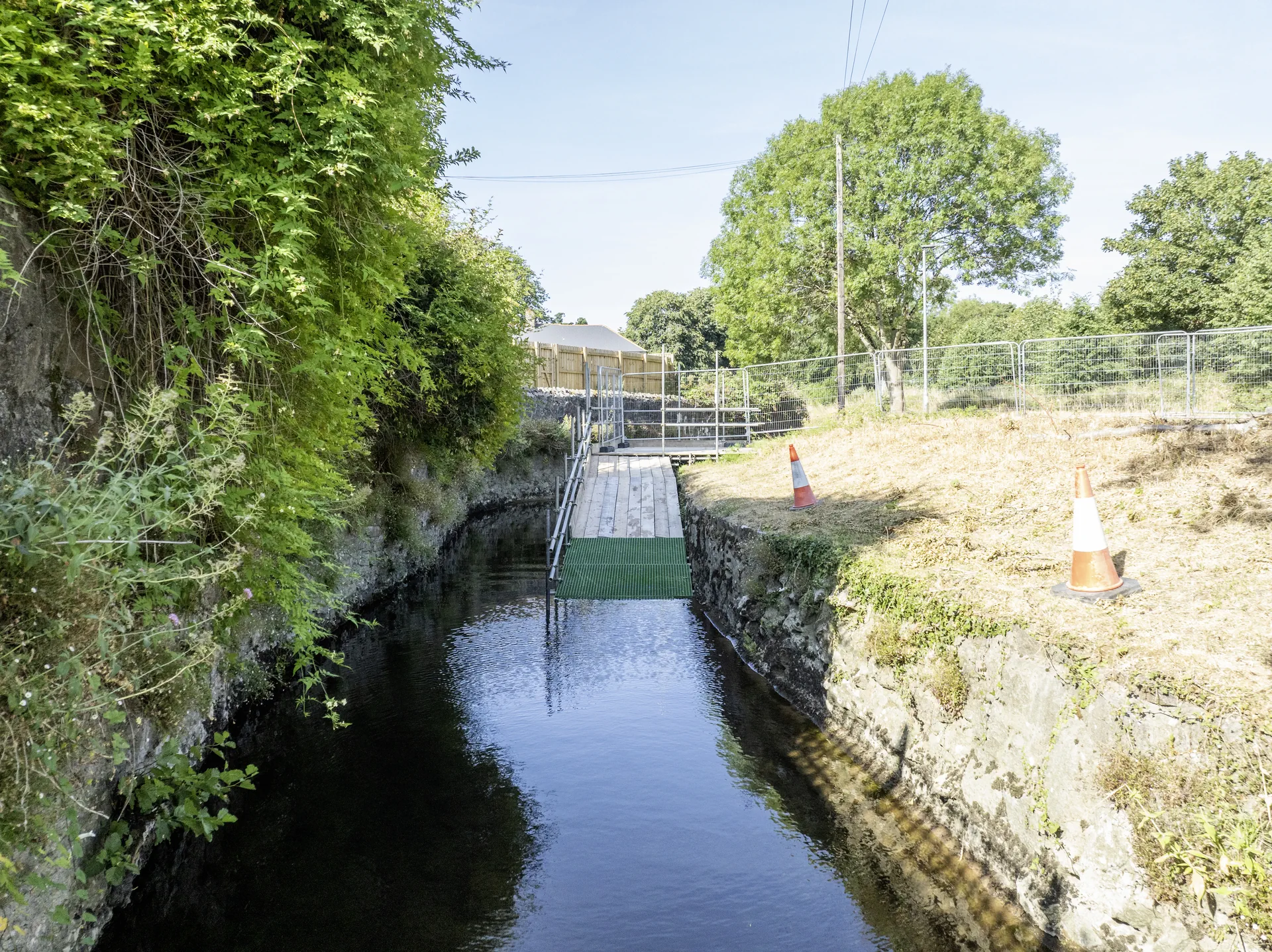 Stone-lined watercourse between vegetated banks, with a temporary access platform, traffic cones and construction fencing beyond