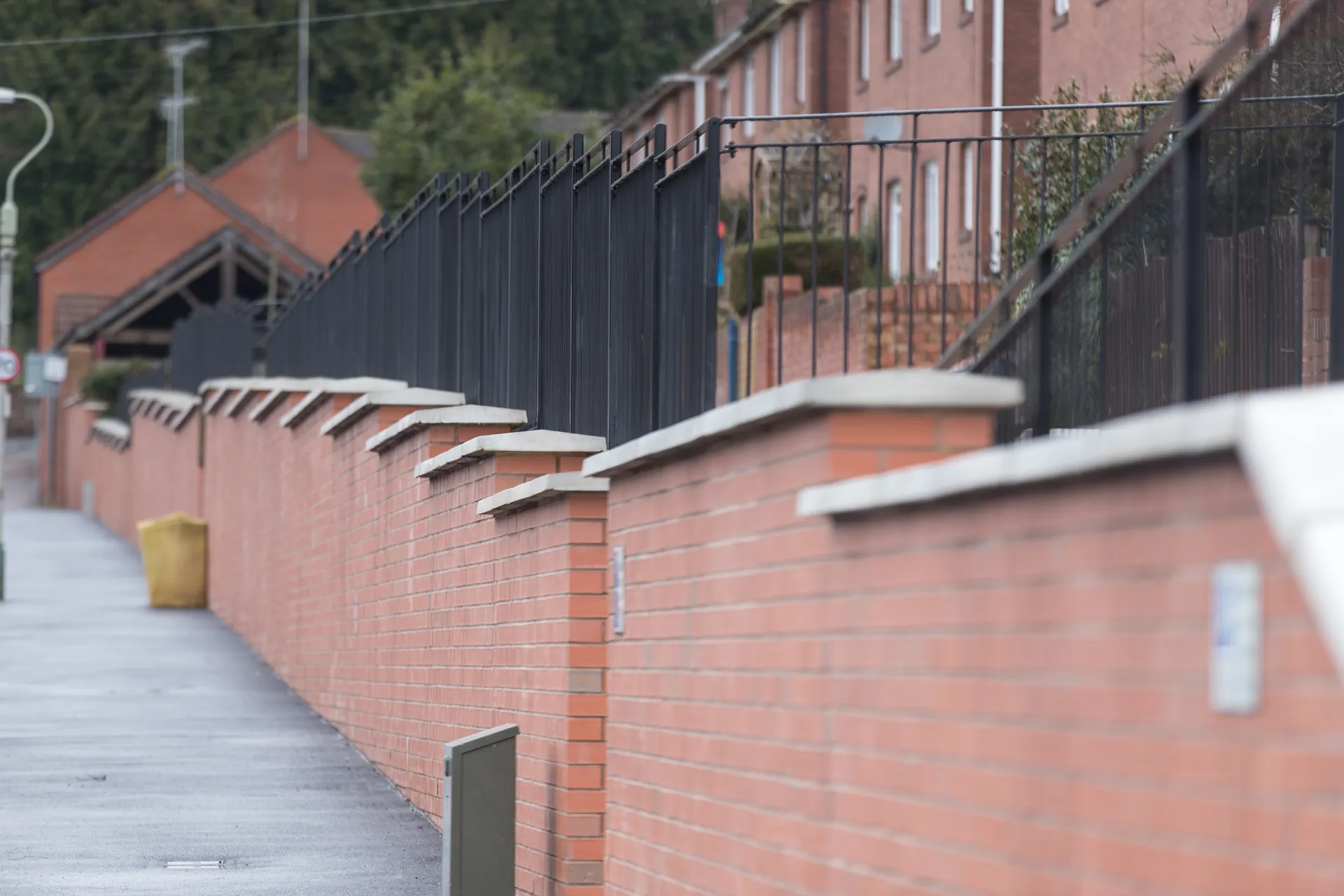 Stepped red-brick wall with concrete copings and metal railings beside a footpath and houses