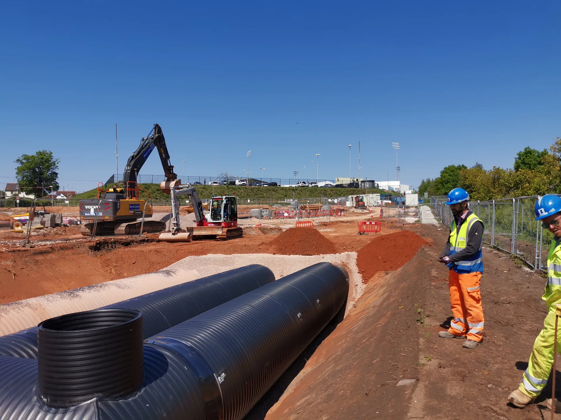 Large black twin-wall pipes beside a trench, with operatives in hi-vis and an excavator on site